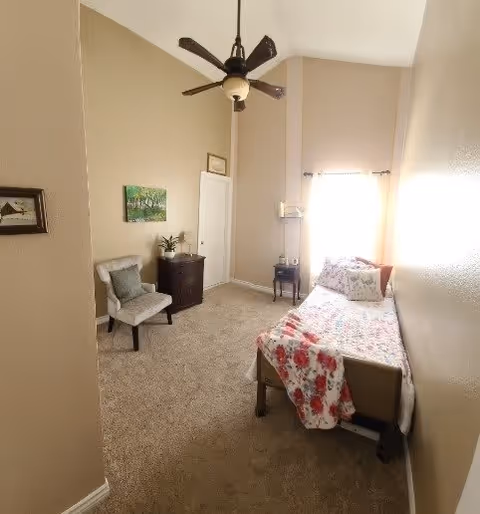 A small bedroom with beige walls and carpeted floor. There is a single bed with floral bedding positioned against the right wall near a window with sheer curtains. A ceiling fan with a light hangs from the ceiling. On the left side of the room, there is a cushioned chair, a small dark wooden dresser with a plant on top, and framed artwork on the walls.