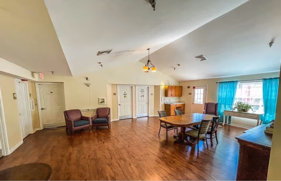 A spacious common area with wooden flooring featuring a wooden dining table with four chairs, two armchairs against the wall, a small cabinet with a sink, and a window with blue curtains letting in natural light.