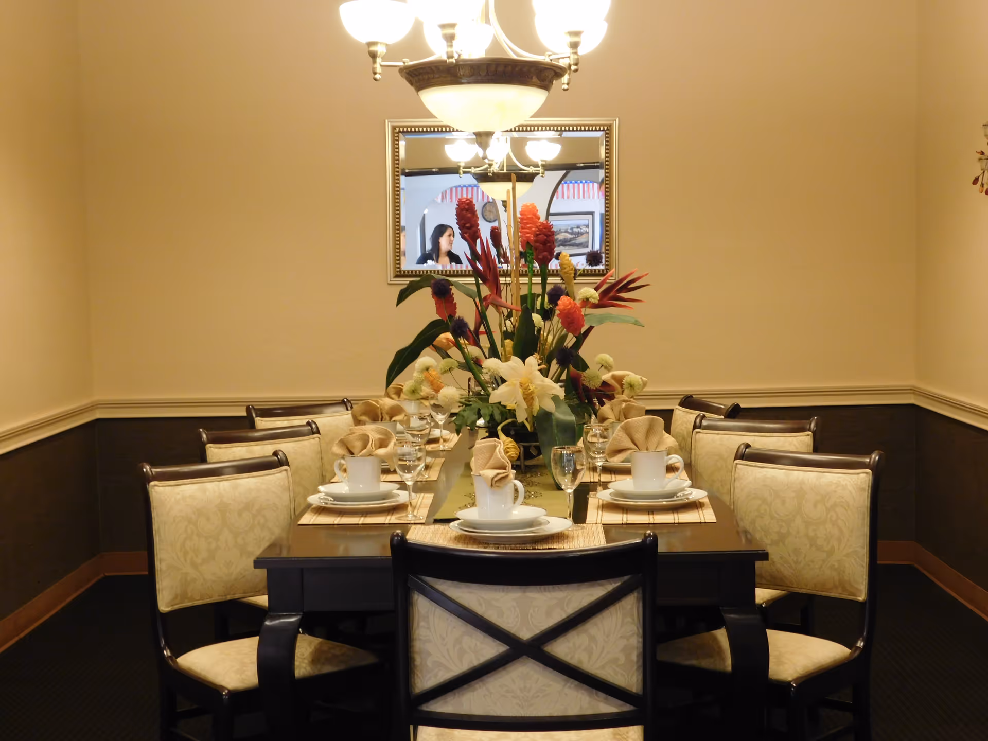 A formal dining room with a rectangular wooden table set for eight people. The table is decorated with a large floral centerpiece and place settings including plates, cups, glasses, and folded napkins. The room has beige walls with a dark brown wainscoting and a chandelier hanging above the table. A mirror on the wall reflects part of the room and a person in the background.