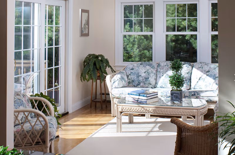 A bright living room with large windows showing green trees outside. The room features a floral-patterned sofa and matching armchair, a glass-top coffee table with books and a small potted plant, a wooden plant stand with a fern, and a wicker chair. Sunlight streams through the windows and glass door, illuminating the hardwood floor and light-colored walls.