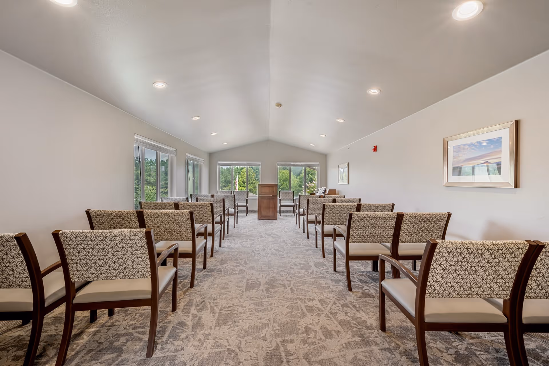 A bright, spacious room with rows of patterned chairs facing a wooden podium at the front. Large windows on the left and at the far end let in natural light and offer views of greenery outside. The room has a light-colored carpet and white walls with framed artwork.