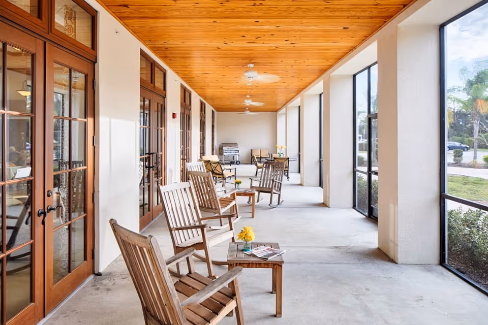 A covered outdoor patio area with wooden rocking chairs and small tables arranged along the side. The ceiling is wooden with ceiling fans, and large glass doors line one side while screened windows overlook a landscaped outdoor area with greenery and palm trees.