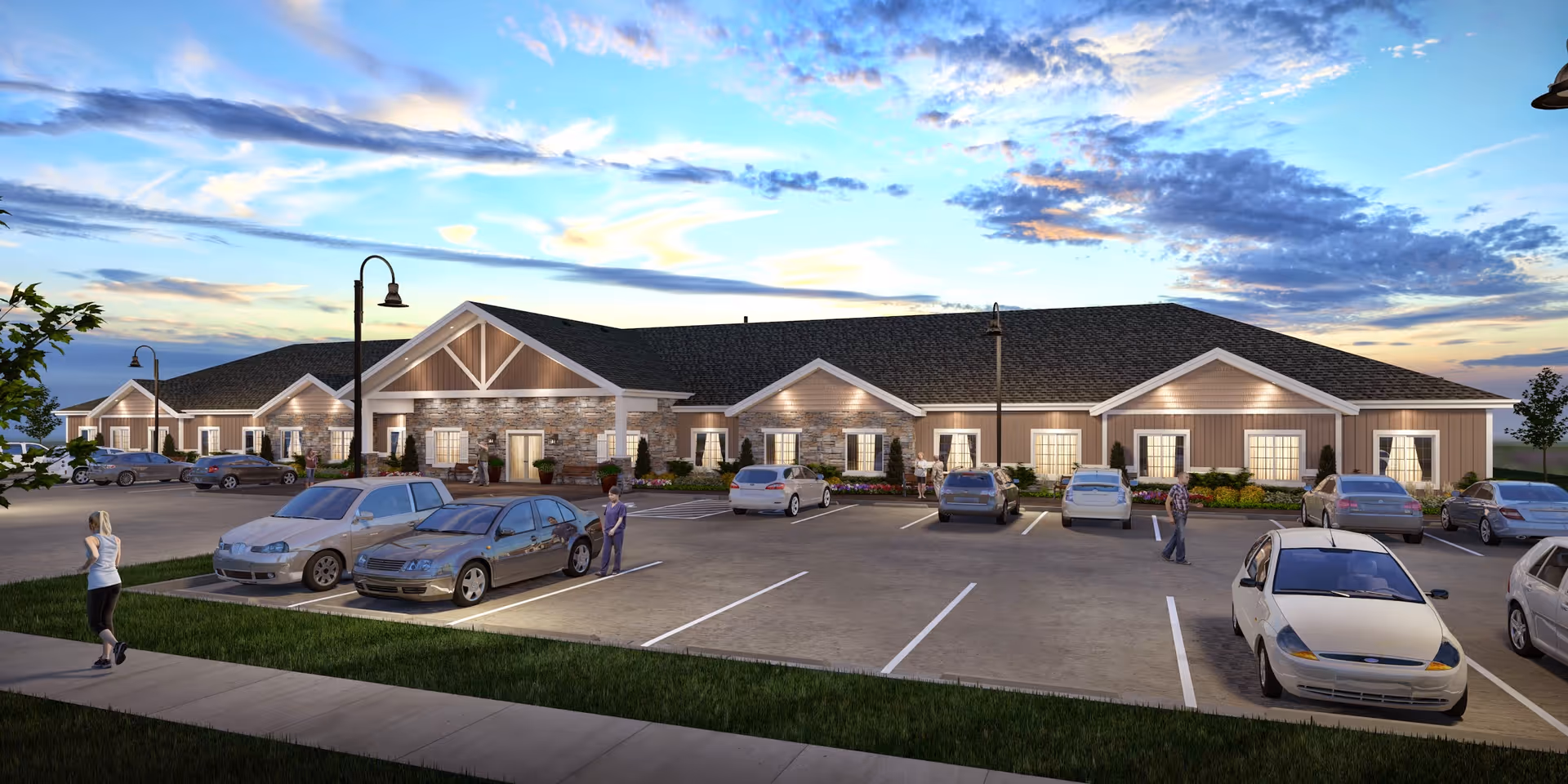 Exterior view of The Legacy Assisted Living facility at dusk, showing a single-story building with stone and siding facade, multiple windows with curtains, and a parking lot with several cars and a few people walking nearby under a partly cloudy sky.