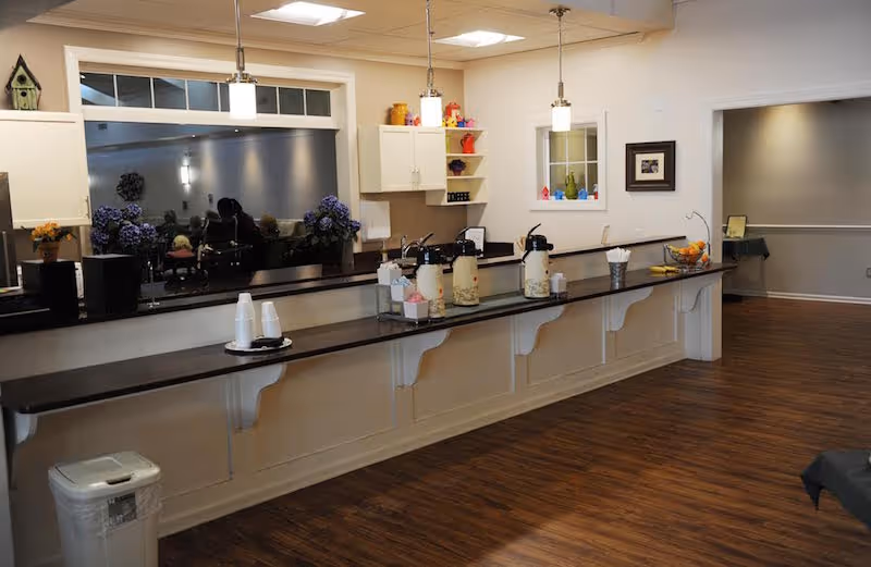 Long interior serving counter with coffee urns, cups, pendant lights and decorative shelves in a communal area.
