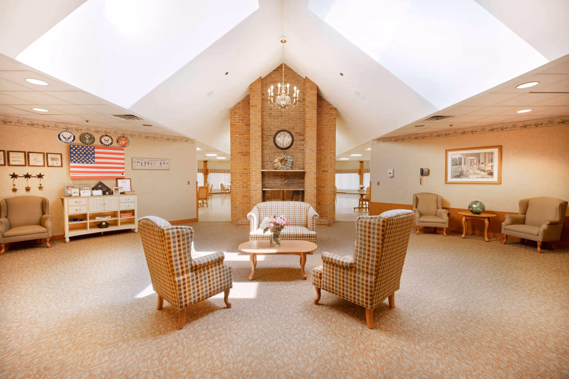 A spacious, well-lit common area with a vaulted ceiling and skylights. The room features a brick fireplace with a clock and decorative wreath above it. There are four plaid armchairs arranged around a wooden coffee table with a vase of flowers. Additional seating includes two beige armchairs and a small wooden table with a decorative globe. The walls are adorned with framed pictures, an American flag, and military insignia. The carpet is light-colored with a subtle pattern.