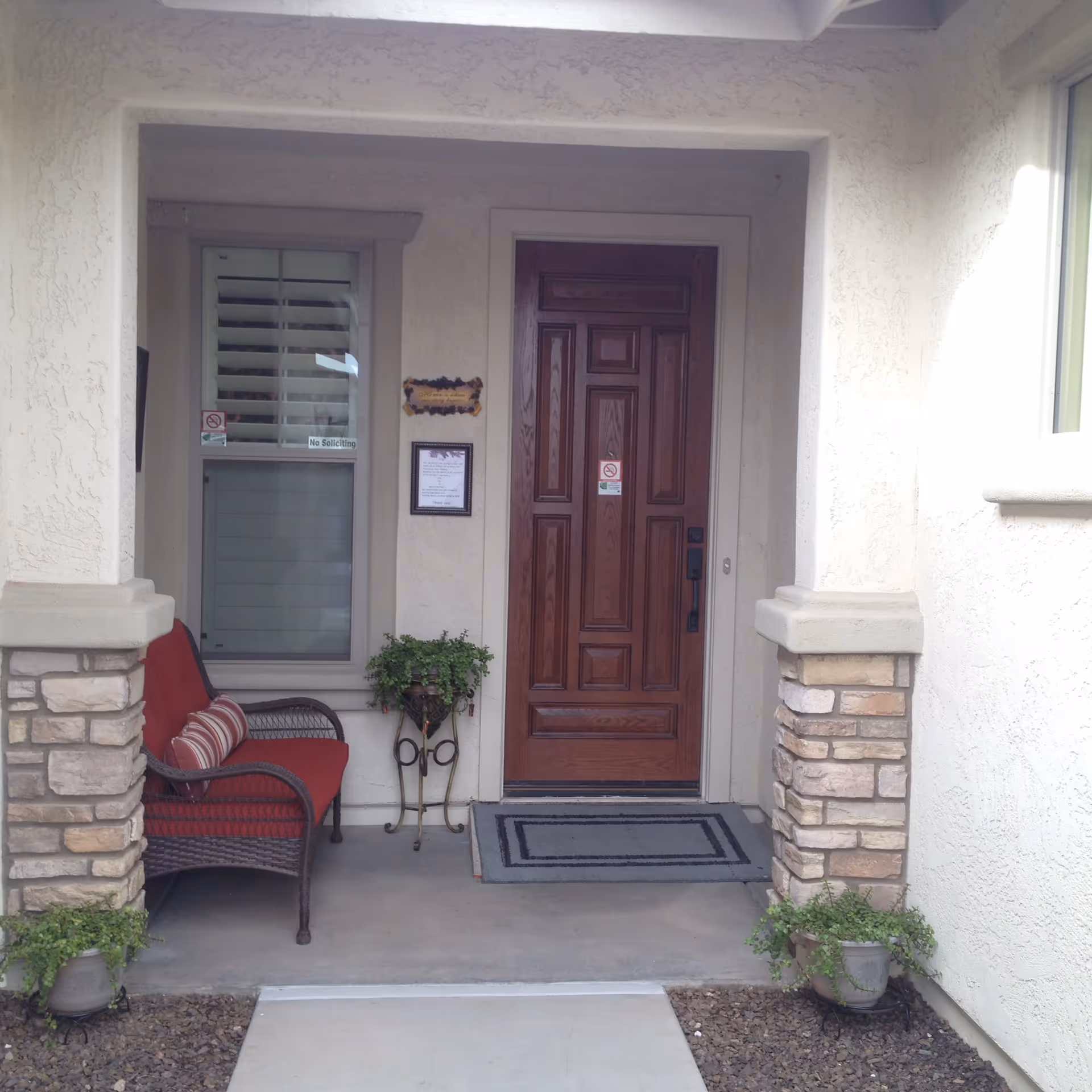 Front porch area of a home with a wooden door, a red cushioned bench, potted plants, and a welcome sign on the wall next to the door.