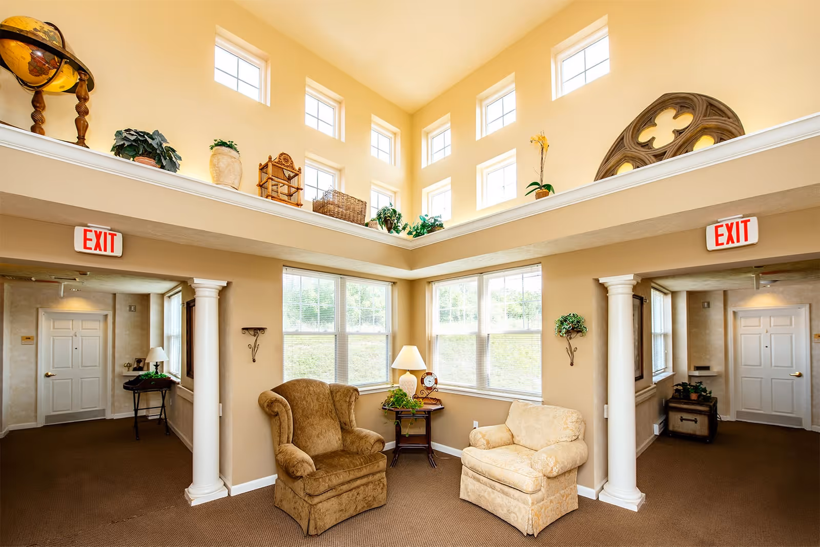 Sunlit two-story common room with upholstered armchairs, tall windows, decorative columns and potted plants.