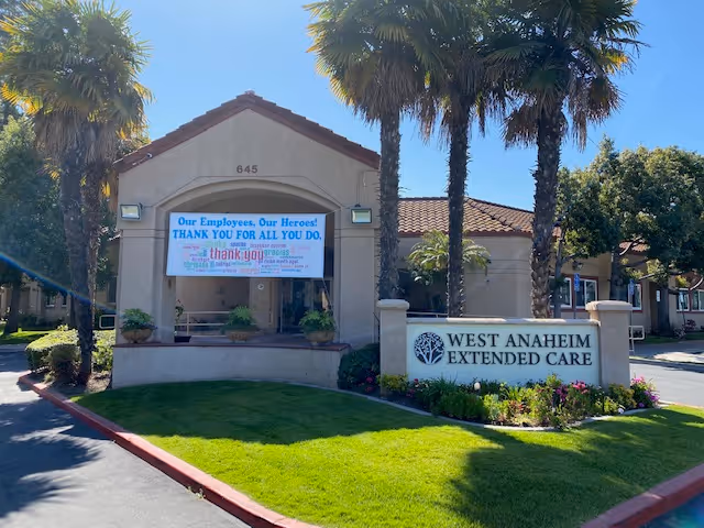 Exterior view of a building with a sign that reads 'WEST ANAHEIM EXTENDED CARE' surrounded by palm trees and landscaping. A banner on the building says 'Our Employees, Our Heroes! THANK YOU FOR ALL YOU DO.' The building has a beige stucco finish with a red tile roof and the number 645 above the entrance.