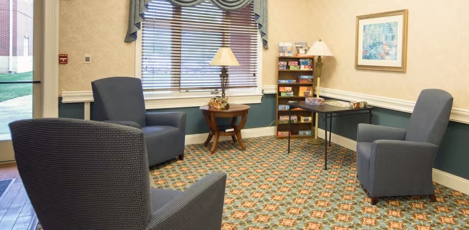 A cozy sitting area with three blue upholstered armchairs arranged around a small wooden table with a lamp. There is a window with blinds and a valance, a framed picture on the wall, and a shelving unit filled with board games next to a floor lamp. The room has patterned carpet and light-colored walls with a dark green wainscoting.