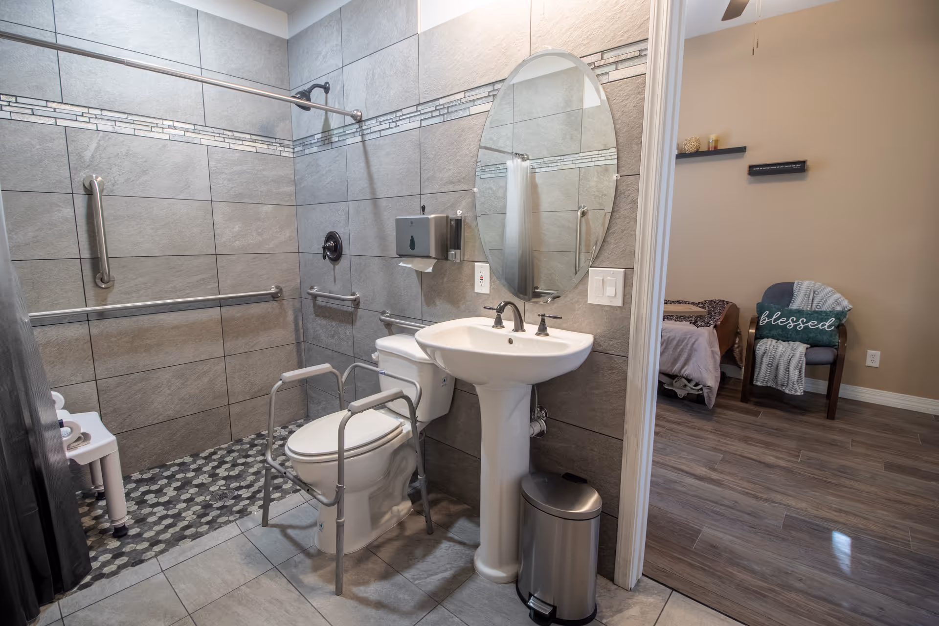A bathroom with gray tiled walls and floor, featuring a walk-in shower with grab bars, a white toilet with safety rails, a pedestal sink with a round mirror above it, and a small trash can. The bathroom opens into a room with wooden flooring, a chair with a blanket and a pillow that says 'blessed', and a bed partially visible.