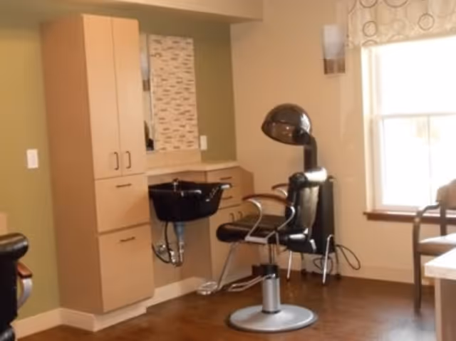 A salon-style room with a styling chair, hooded hair dryer, shampoo basin, and storage cabinets next to a window.