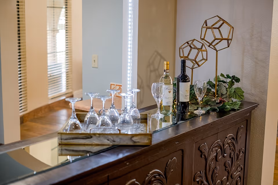 Decorative wooden console topped with wine bottles, multiple wine glasses, geometric metal sculptures and greenery in an interior common area.