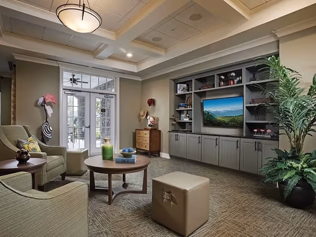 A cozy living room area with two green armchairs, a round wooden coffee table, a beige ottoman, and built-in shelves with a flat-screen TV displaying a scenic mountain view. The room has a carpeted floor, a potted plant, and a glass door leading outside. Hats and decorations are hung on the walls.