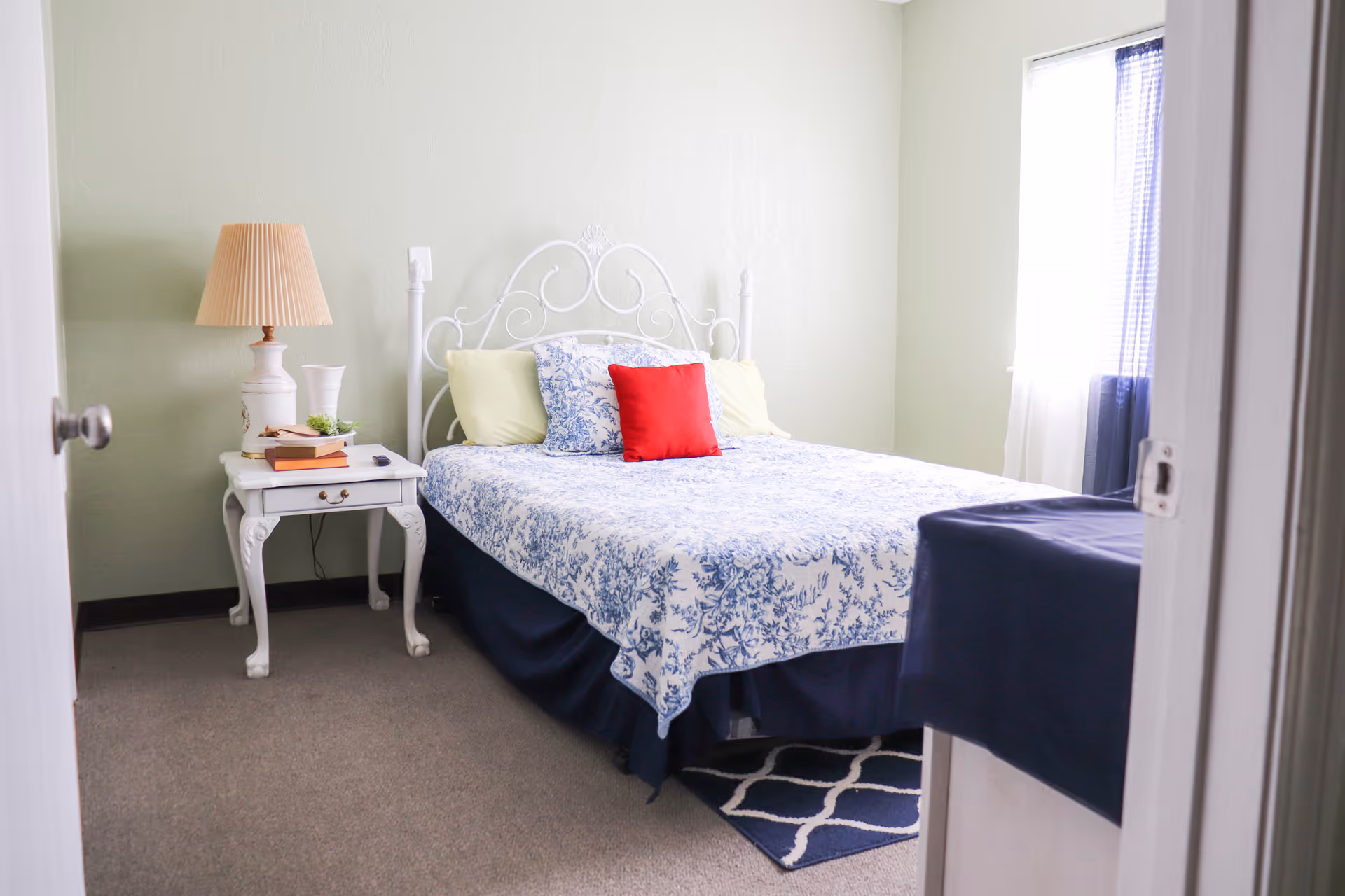 Sunlit bedroom with a white metal bed dressed in blue floral bedding, a red accent pillow, and a white nightstand with a lamp.