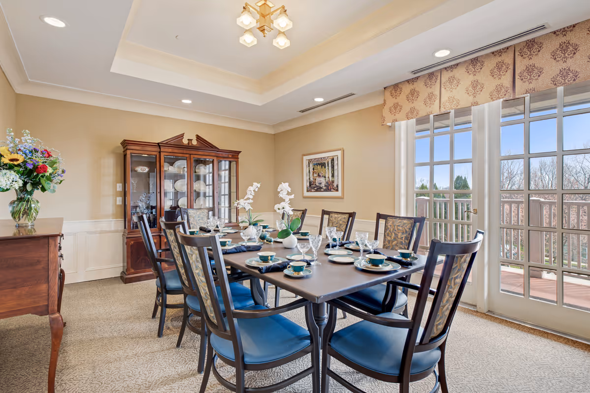 A well-lit dining room with a rectangular table set for six people with plates, cups, and glassware. The chairs have blue cushions and patterned backs. There is a wooden china cabinet filled with dishes against the wall, a sideboard with a vase of flowers, and large windows with patterned valances letting in natural light and showing an outdoor view.