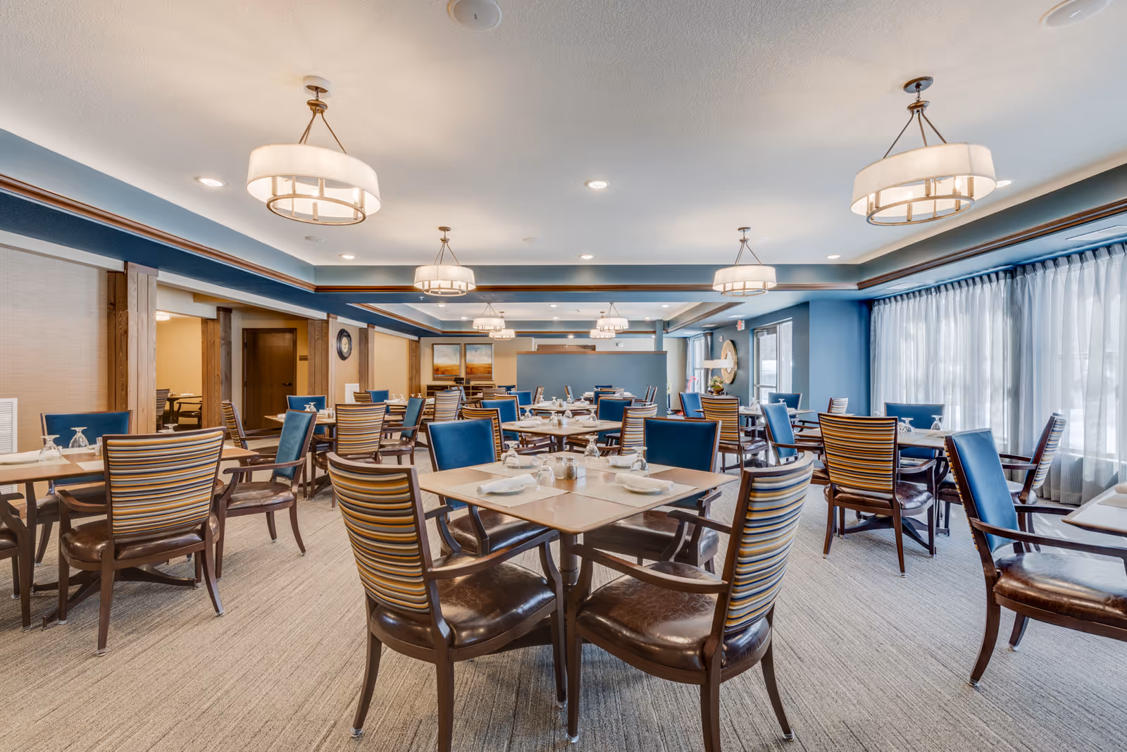 Spacious dining room with multiple set tables and chairs, large windows with curtains, and ceiling light fixtures.