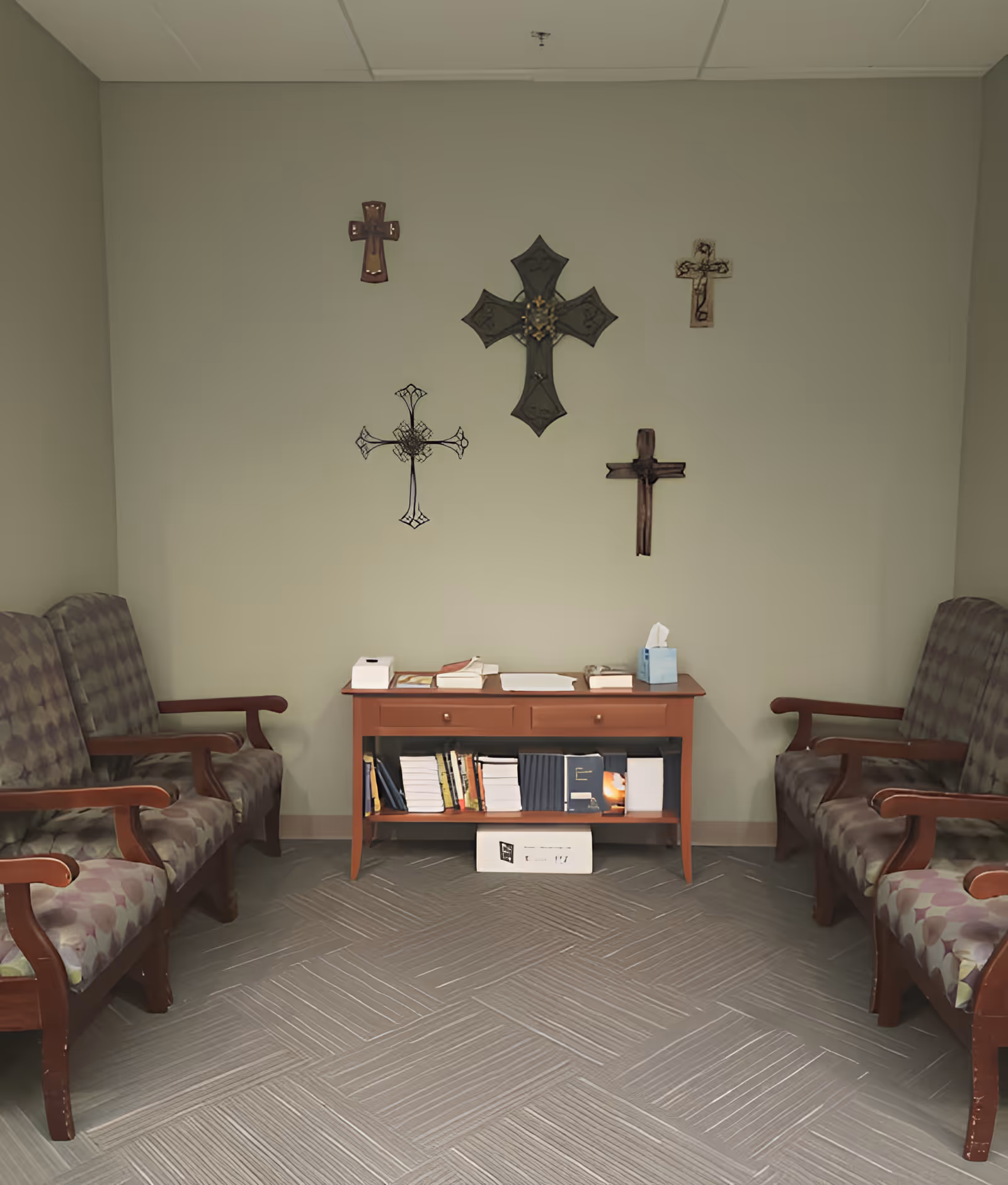 A small room with two patterned cushioned wooden benches facing each other. Between them is a wooden table with books, tissues, and other items on top and shelves below filled with more books. Five decorative crosses are mounted on the light green wall above the table.
