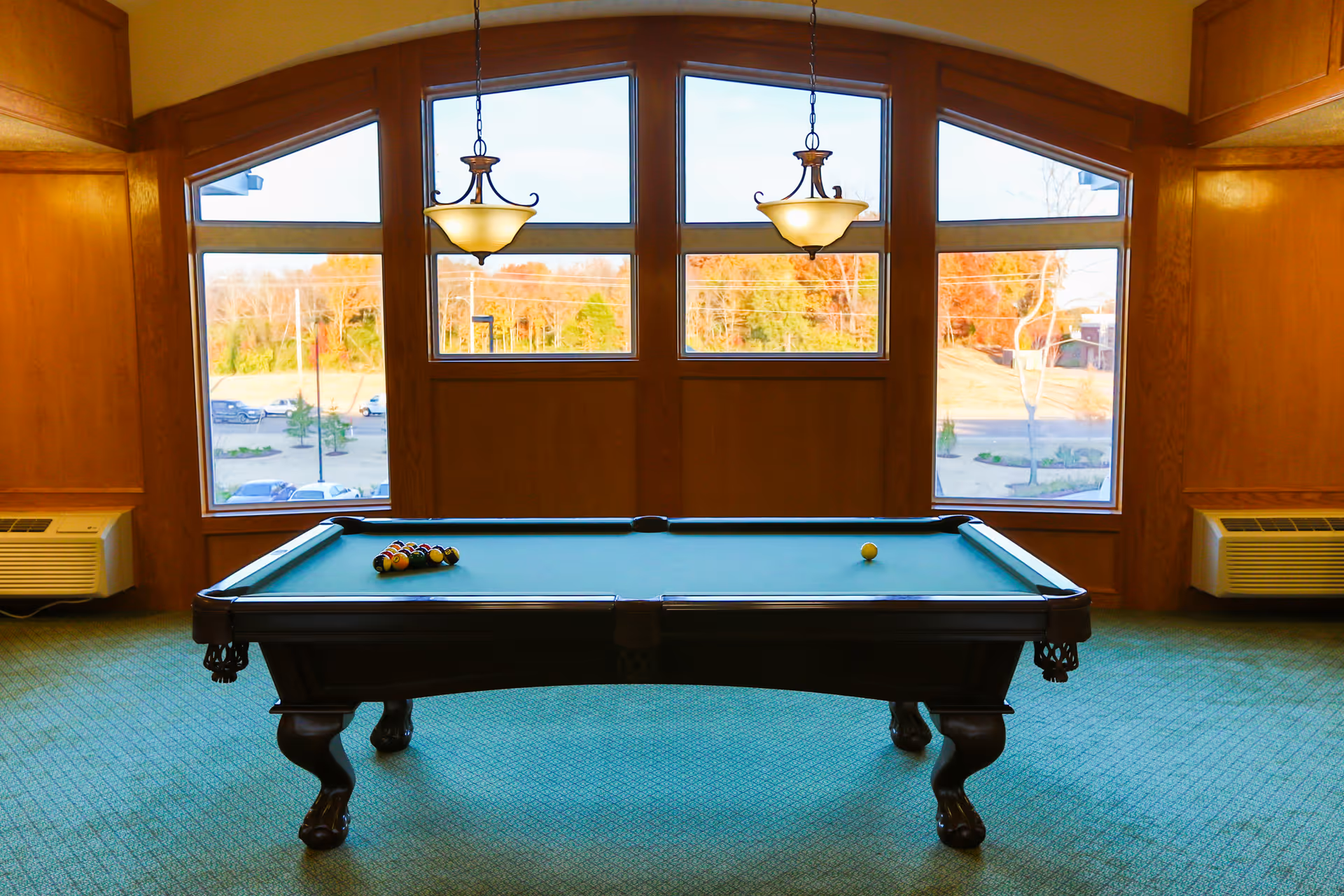 A pool table centered in a wood-paneled room with large arched windows and two hanging light fixtures.