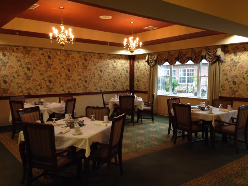 A dining room with several tables covered with white tablecloths, each set with cups, glasses, and napkins. The room has floral wallpaper, a large window with draped curtains, and two chandeliers hanging from the ceiling providing warm lighting.