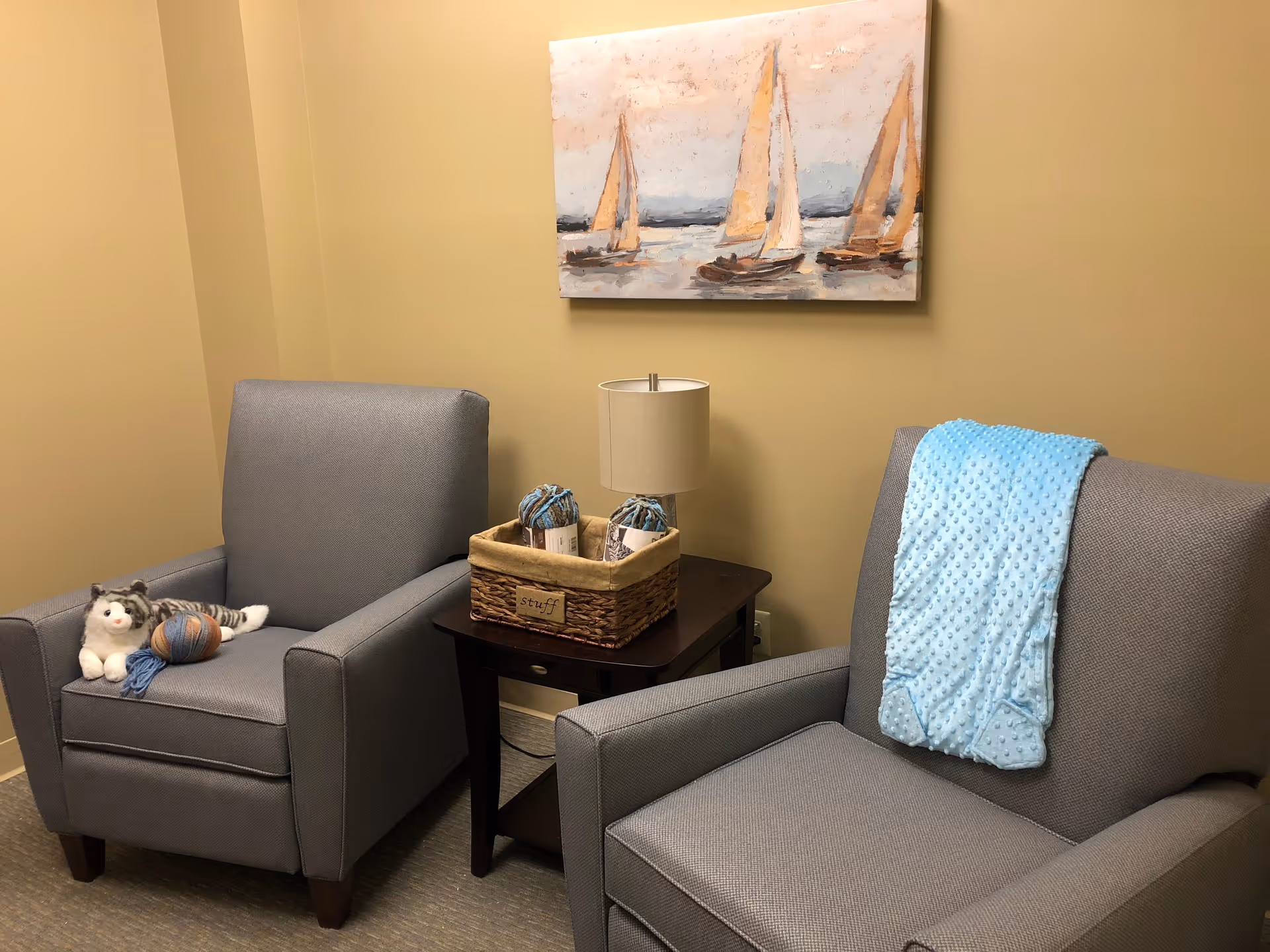 A cozy seating area with two gray armchairs separated by a dark wooden side table. One chair has a stuffed cat toy and a ball of yarn on it, while the other chair has a light blue textured blanket draped over the back. On the side table, there is a basket labeled 'stuff' containing two balls of yarn and a small lamp. A painting of sailboats on water hangs on the beige wall behind the chairs.