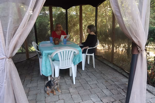 Two women sit at a table covered with a teal tablecloth inside a screened gazebo while a small dog rests on the stone pavers.
