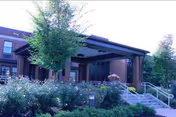 Exterior view of a brick building with a covered entrance, stairs with handrails, and landscaped greenery including bushes and trees in front.
