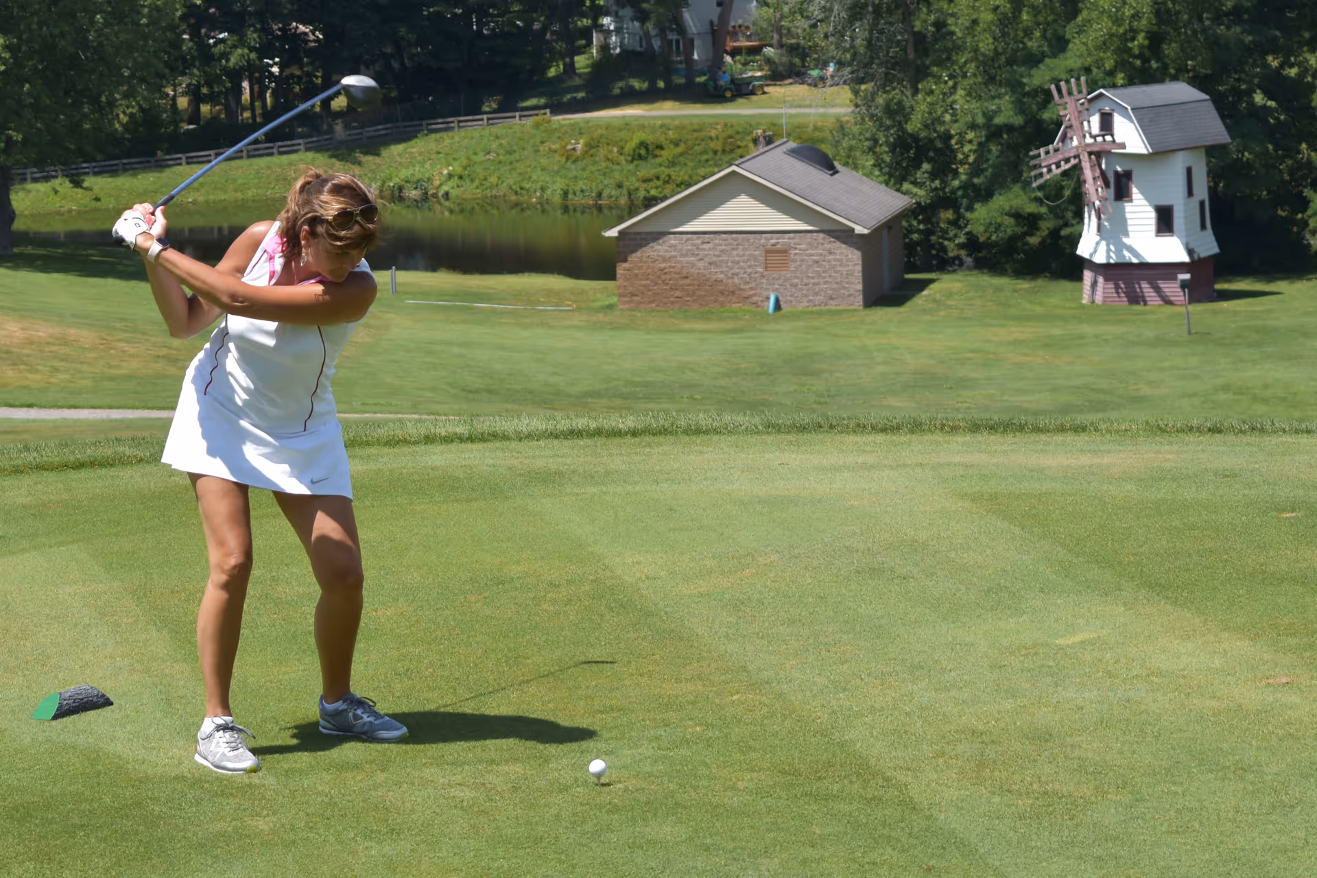 A woman in a white golf outfit is swinging a golf club on a green golf course. In the background, there is a small building and a decorative windmill structure, with trees and a pond further behind.