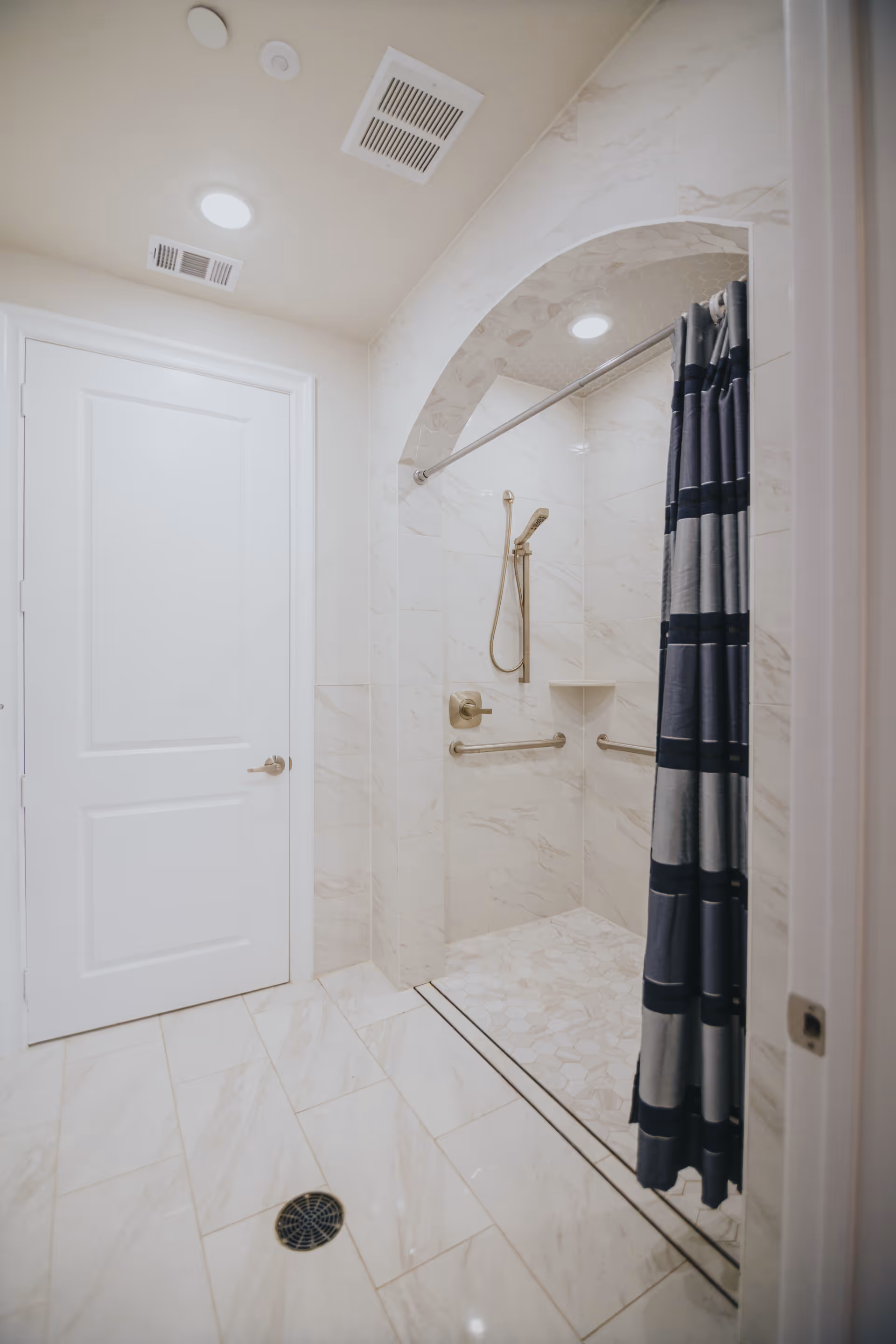 A clean, modern bathroom with a walk-in shower featuring a curved arch entrance, beige marble tiles, a handheld showerhead, grab bars, and a dark blue and gray striped shower curtain. There is a white door and ceiling vents visible.