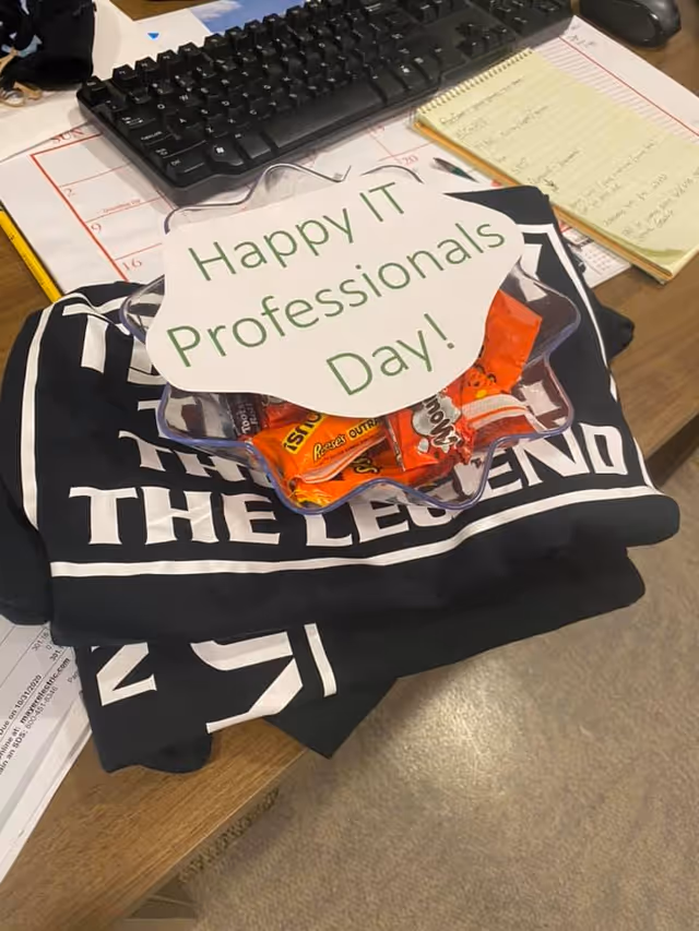 A desk with a keyboard, notepad, folded shirts and a bowl of candy topped with a sign reading "Happy IT Professionals Day!"