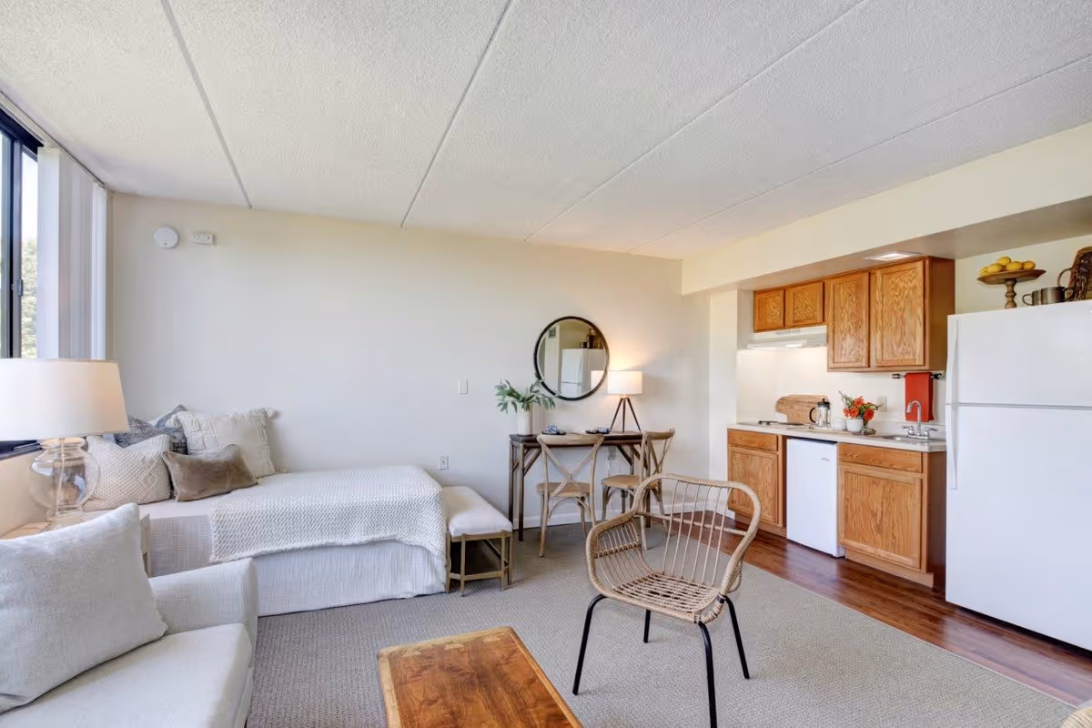A bright studio apartment featuring a bed with multiple pillows and a throw blanket next to a window with a lamp on a side table. There is a small wooden table with two chairs and a round mirror on the wall. The kitchenette includes wooden cabinets, a white refrigerator, a dishwasher, a stove, and a sink. A wicker chair and a wooden coffee table are in the foreground on a carpeted floor.