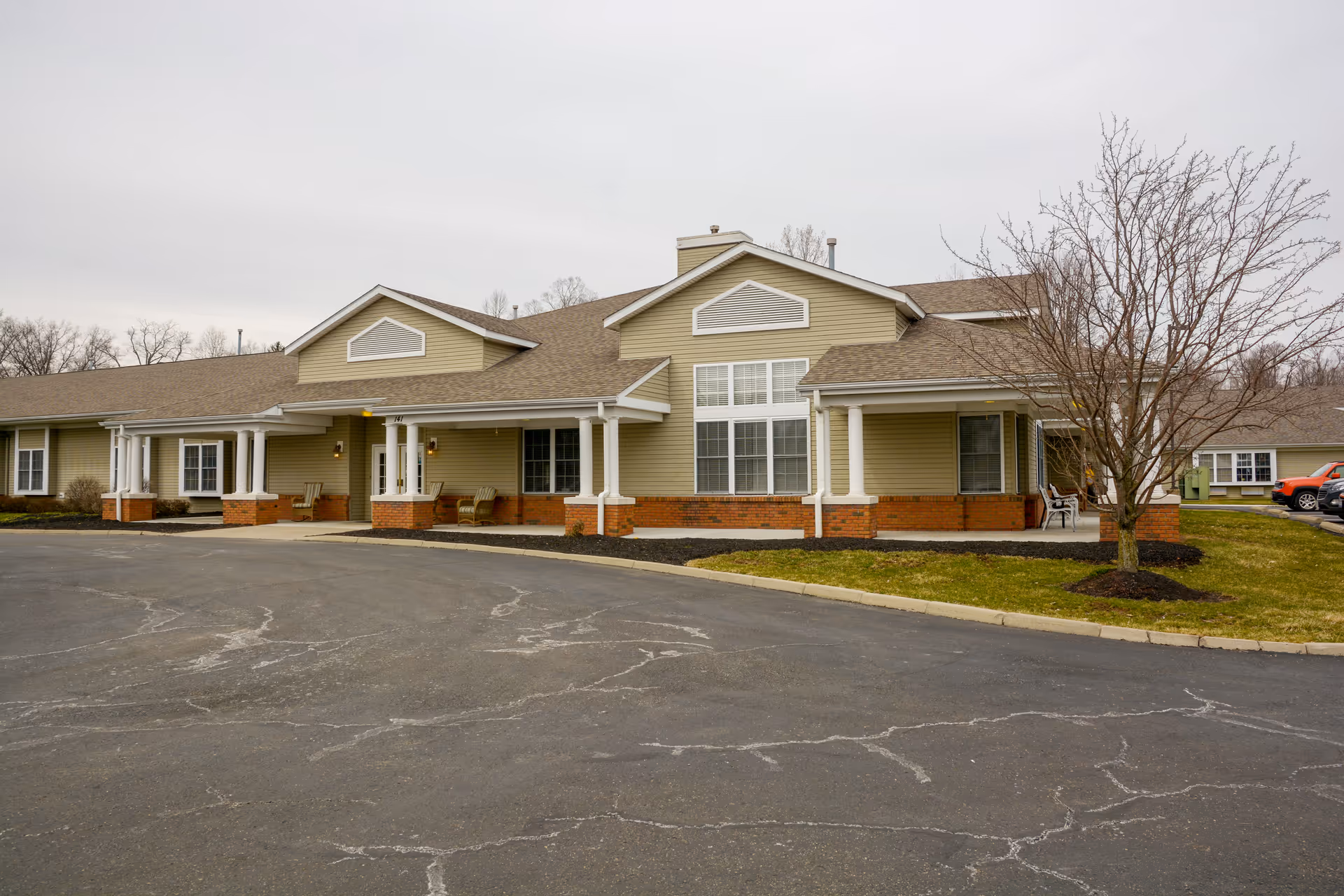 Front exterior of a single-story beige senior living building with columns, a circular driveway, and leafless trees.