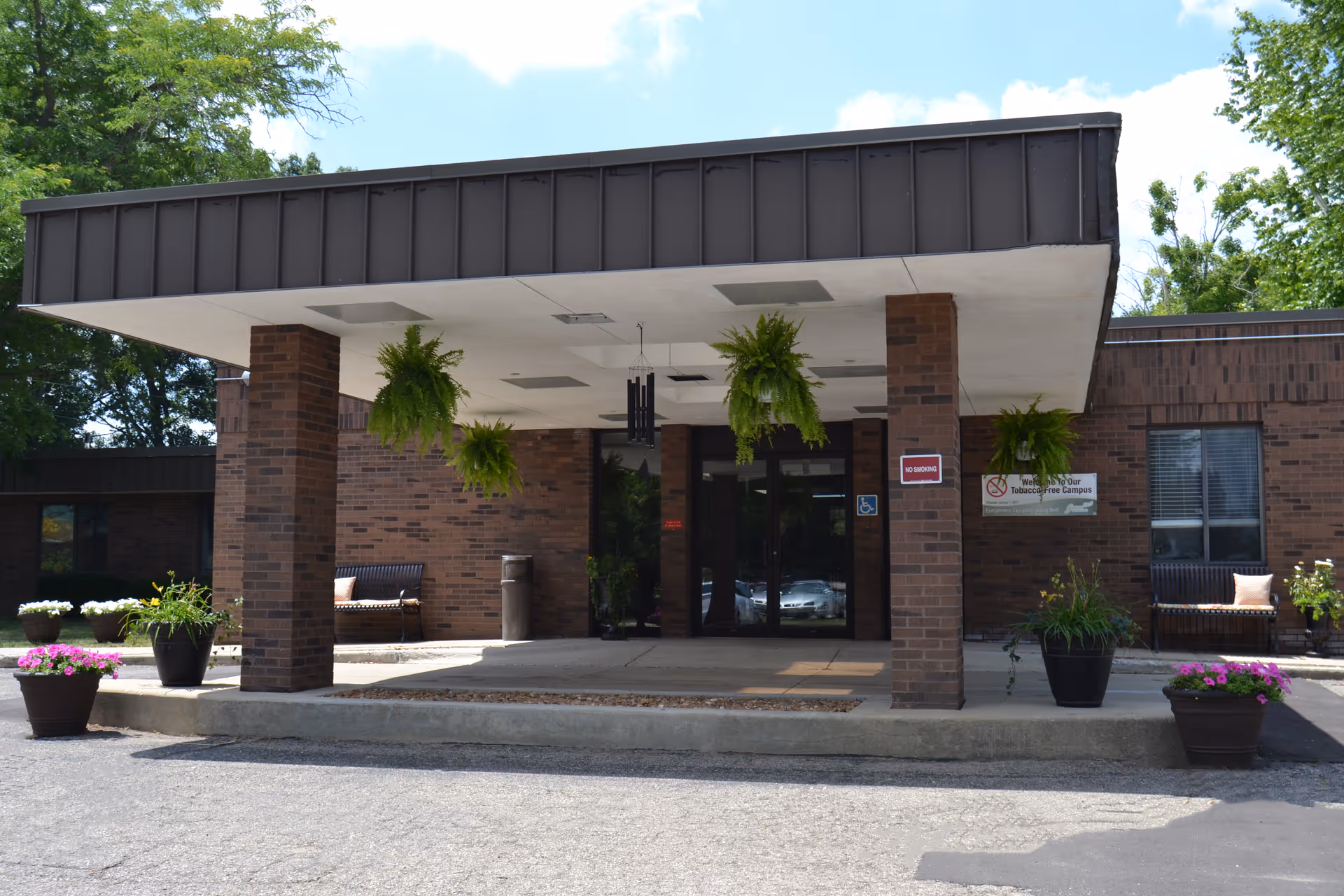 Entrance of a brick building with a covered drop-off area supported by brick columns. Hanging green plants are suspended from the ceiling of the covered area. There are potted plants with flowers on either side of the entrance, benches with cushions, and a sign indicating a tobacco-free campus. The sky is partly cloudy and trees are visible in the background.