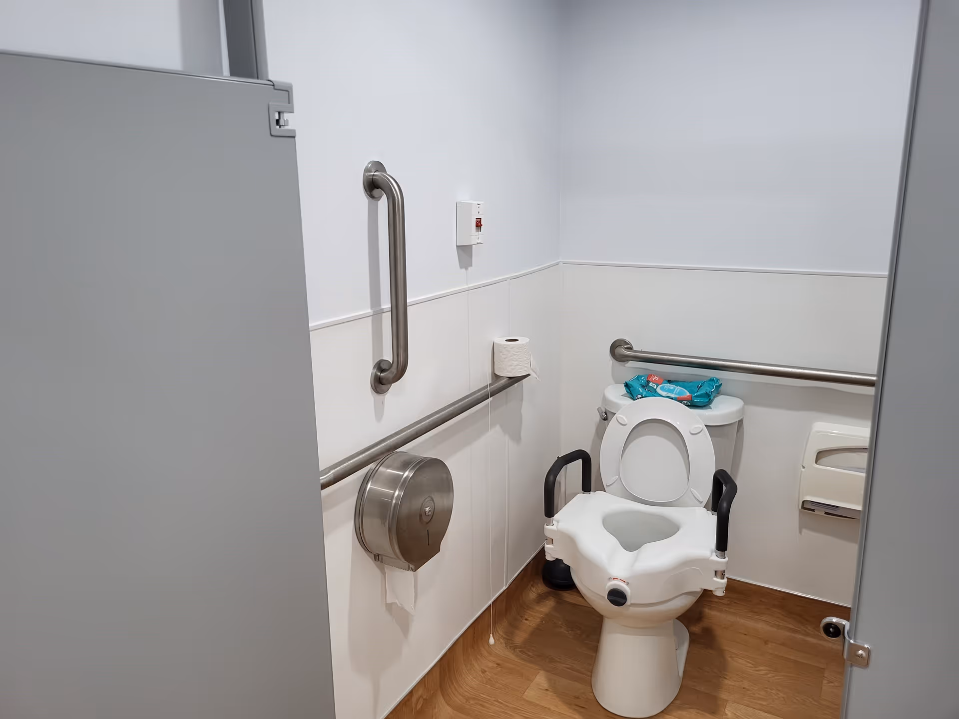 A small accessible bathroom stall with a toilet equipped with armrests for support. There are metal grab bars on the walls, a toilet paper dispenser, a roll of toilet paper on a grab bar, and a pack of wipes on top of the toilet tank. The floor has a wood-like finish and the walls are painted white.