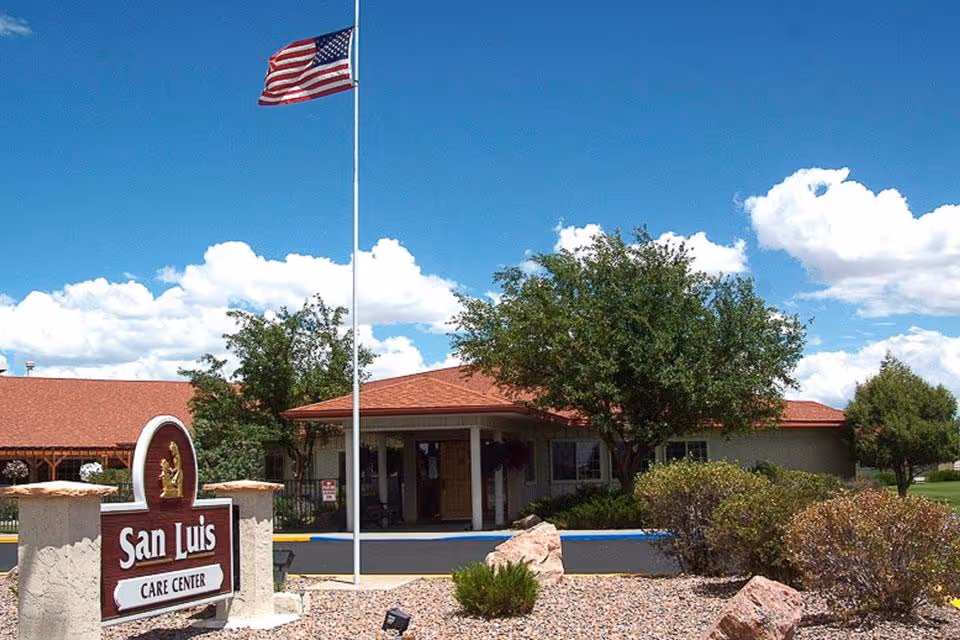 Exterior view of San Luis Care Center building with a red roof, surrounded by trees and bushes under a blue sky with scattered clouds. An American flag flies on a flagpole near the entrance, and a sign in front reads 'San Luis Care Center.'