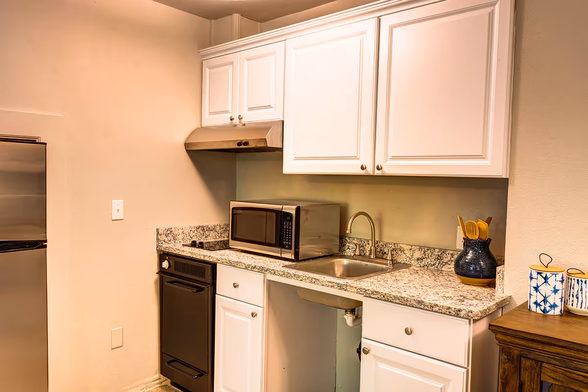 A small kitchen area with white cabinets, a granite countertop, a stainless steel sink with a faucet, a microwave oven, a black stove with an exhaust hood, and a stainless steel refrigerator. On the right side of the countertop, there is a dark blue vase holding wooden utensils and two decorative containers with blue patterns on a wooden surface.