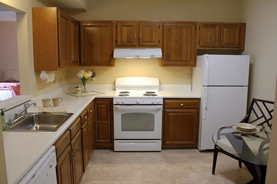 A kitchen with wooden cabinets, a white electric stove with an overhead vent, a white refrigerator, a stainless steel sink with a soap dispenser, and a glass-top dining table with two chairs. There is a small vase with flowers on the countertop and two mugs near the sink. A bedroom is partially visible through an open doorway on the left.