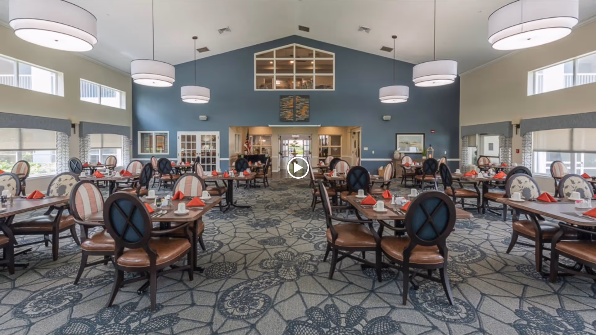 A spacious dining room with multiple tables set with white cups, plates, and red napkins. The room has high ceilings with large round pendant lights, patterned carpet, and windows with shades allowing natural light to enter. The walls are painted blue and beige, and there is a decorative piece above a doorway at the far end of the room.
