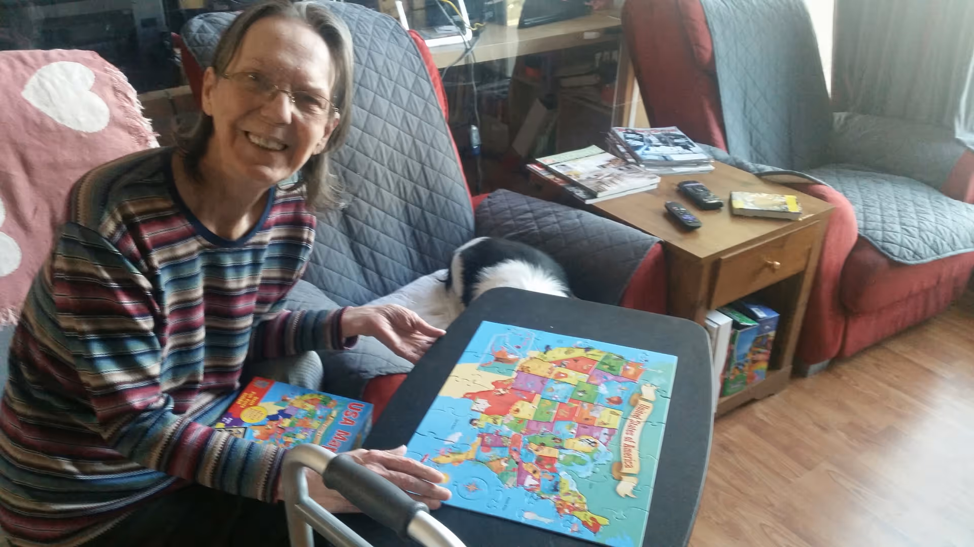 An elderly woman wearing glasses and a striped sweater is sitting on a couch in a living room, smiling while working on a colorful puzzle of the United States on a small black table. The room has two armchairs with quilted covers, a wooden side table with books and remote controls, and a wooden floor.