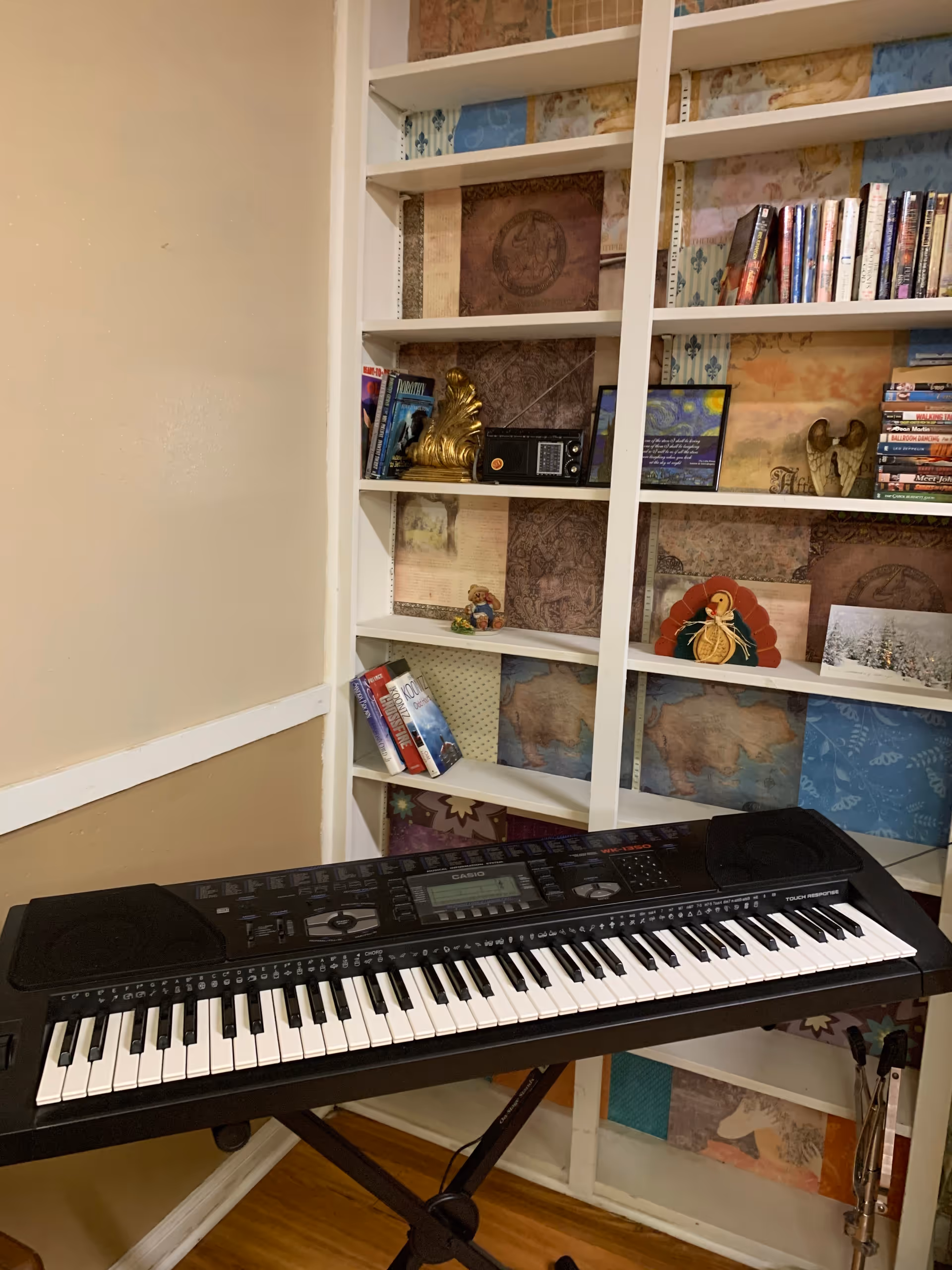 A black Casio electronic keyboard on a stand positioned in front of a white bookshelf filled with books, decorative items, and framed pictures. The wall behind the bookshelf has a patchwork-style wallpaper with various patterns and colors.