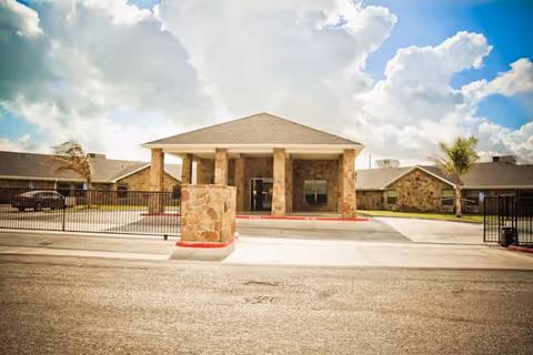 Front exterior view of Windsor Nursing and Rehabilitation Center of Corpus Christi, showing a stone building with a covered entrance, a gated driveway, and a partly cloudy sky above.