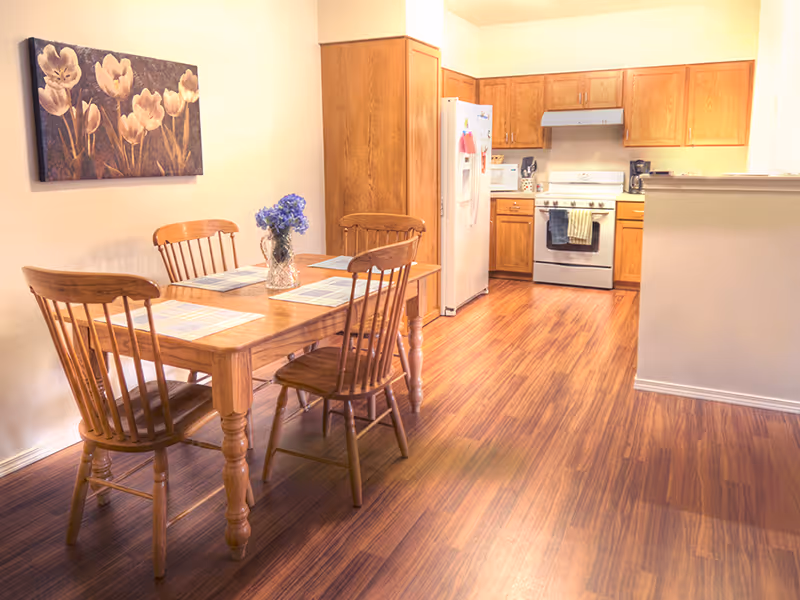 Wood dining table with four chairs and a small vase in an open dining area leading to a kitchen with wooden cabinets and appliances.