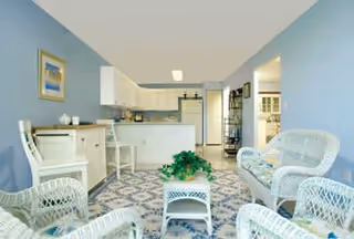 Open-plan living area with white wicker seating and a small coffee table on a patterned rug, facing a kitchen with light blue walls.