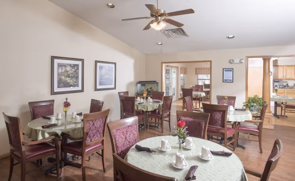 A dining room with several round tables covered with light green tablecloths, each set with white cups, saucers, and brown napkins. The chairs have wooden frames with red upholstered backs and seats. There are framed pictures on the beige walls, a ceiling fan with lights, and an adjacent room visible through open doorways.