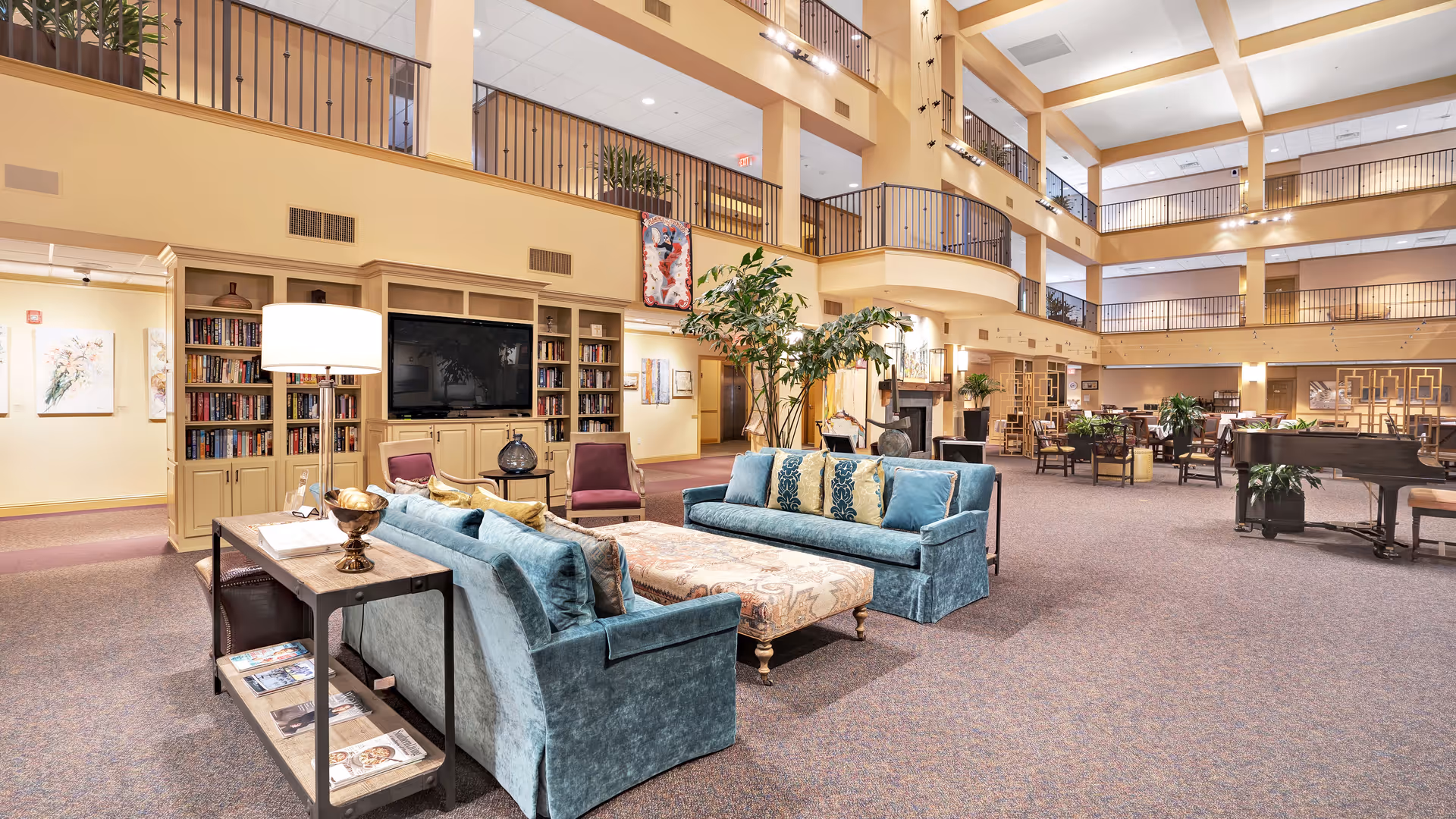 Spacious multi-story retirement community common area with sofas, bookshelves, a TV, plants, and a grand piano.