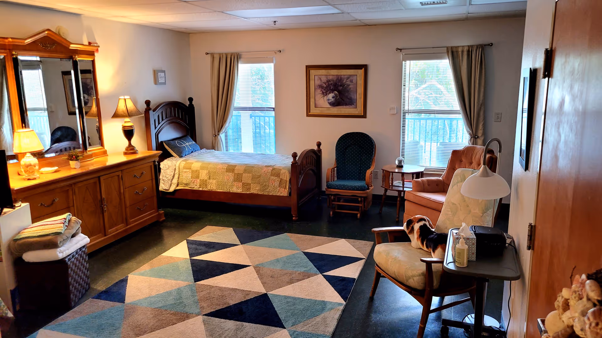 A cozy bedroom in an assisted living facility featuring a wooden bed with a patchwork quilt, a large wooden dresser with a mirror and two lamps, a patterned area rug, two windows with beige curtains, a framed picture on the wall, a green cushioned rocking chair, a peach armchair, and a cushioned chair with a small dog sitting on it next to a side table with a lamp, tissues, and hand sanitizer.