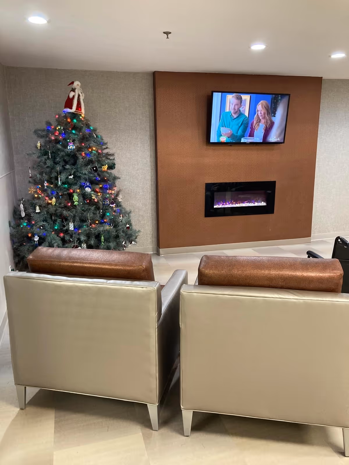 Two armchairs facing a wall-mounted TV and electric fireplace next to a decorated Christmas tree in a lounge.