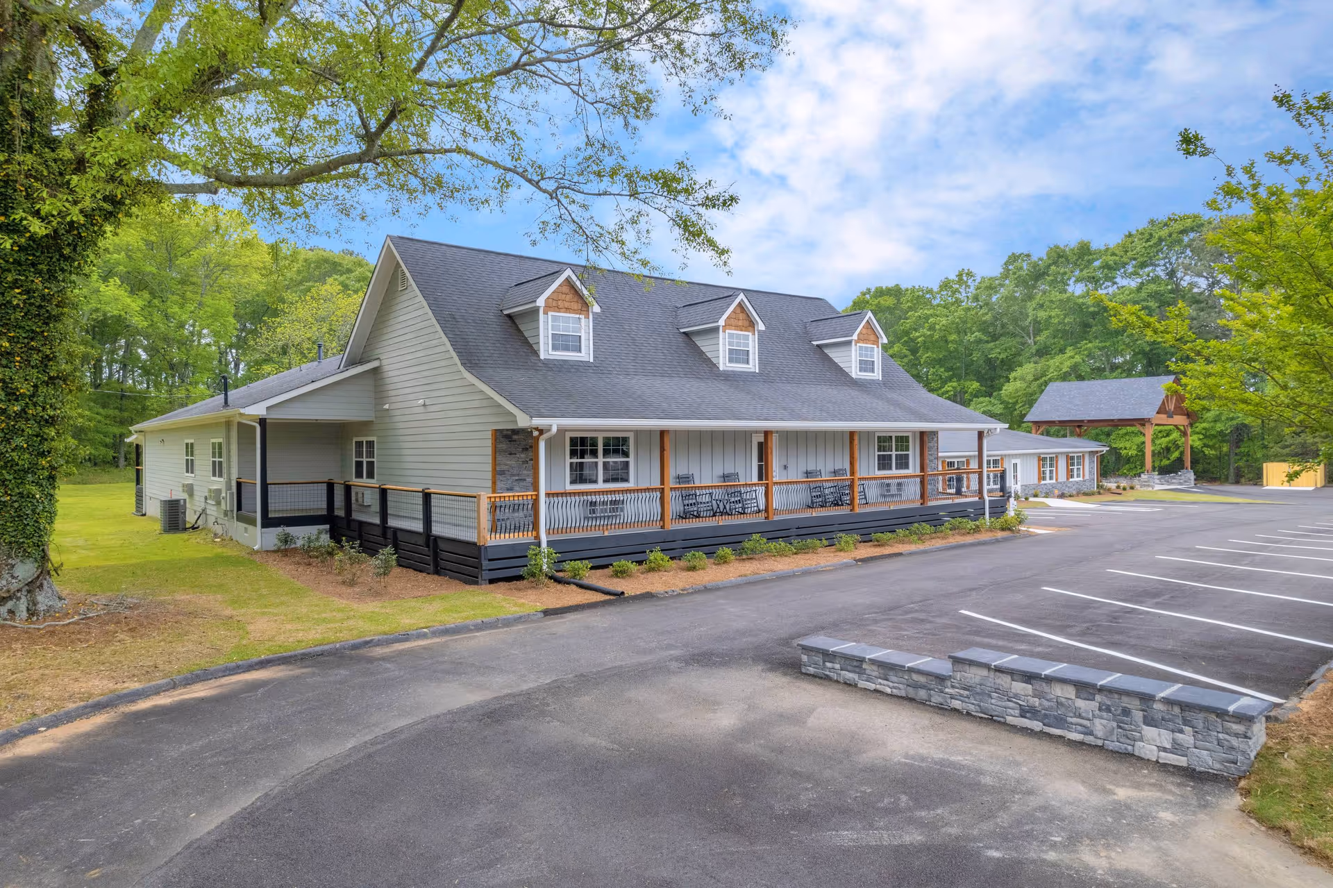A single-story senior living building with a covered front porch, rocking chairs, and an adjacent parking lot surrounded by trees.
