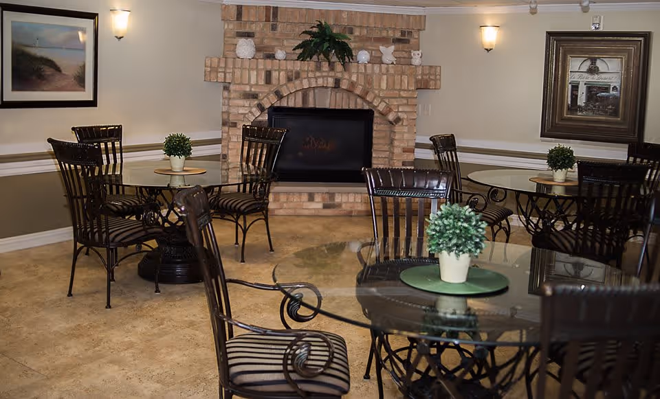 A cozy common area with several round glass-top tables and wrought iron chairs with striped cushions. Each table has a small potted plant centerpiece. The room features a brick fireplace in the center of the back wall, flanked by framed artwork and soft wall lighting.