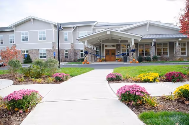 Front exterior view of Bridges by EPOCH at Trumbull, showing a two-story building with a covered entrance decorated with pumpkins and a large blue ribbon. The walkway leading to the entrance is flanked by landscaped flower beds with pink, yellow, and purple flowers and green shrubs.