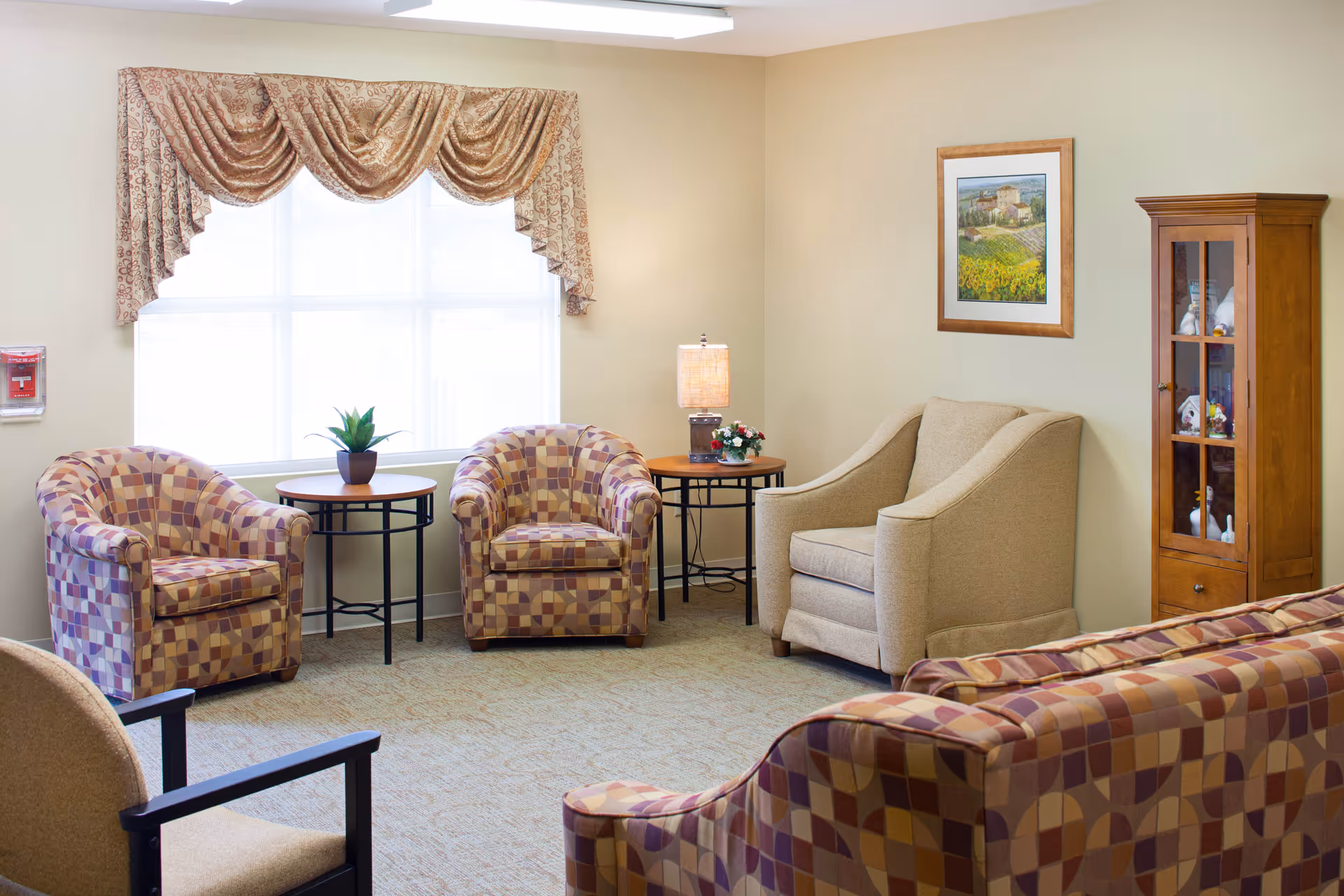 A cozy living room area with patterned armchairs and a matching sofa, a beige armchair, two small round tables with a lamp and a plant, a wooden display cabinet with decorative items, a large window with patterned drapes, and a framed painting on the wall.